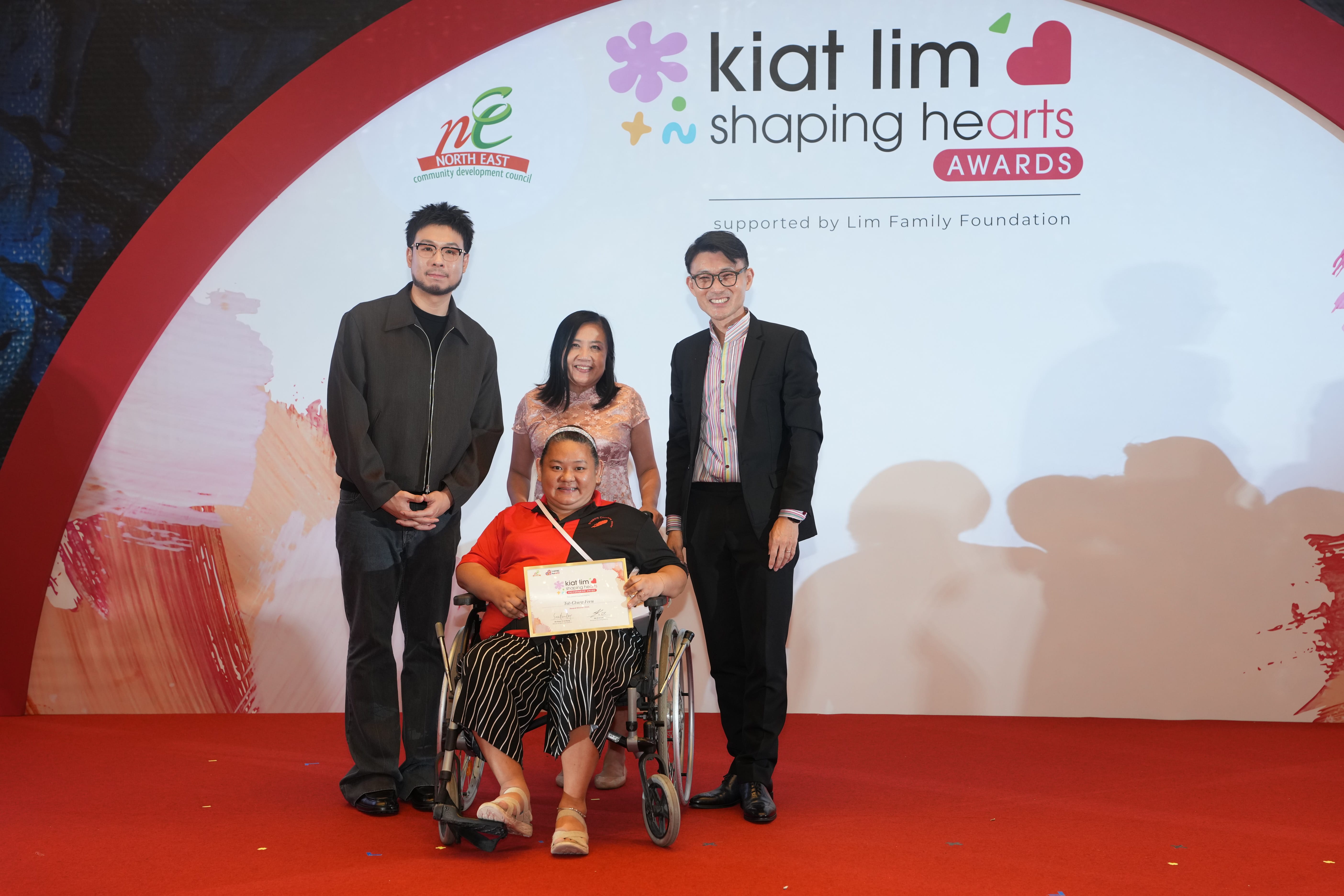 Award recipient standing on stage with the presenter, holding their certificate and posing for a group photo during the award ceremony, with a backdrop reading ‘Kiat Lim-Shaping Hearts Awards'.